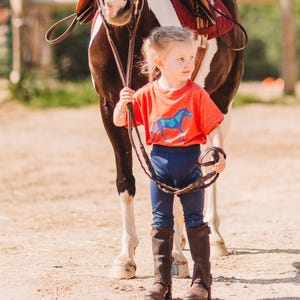 May include: A young child wearing an orange t-shirt with a blue horse graphic, navy blue leggings, and brown riding boots stands next to a horse. The horse has a brown and white coat and is wearing a saddle.