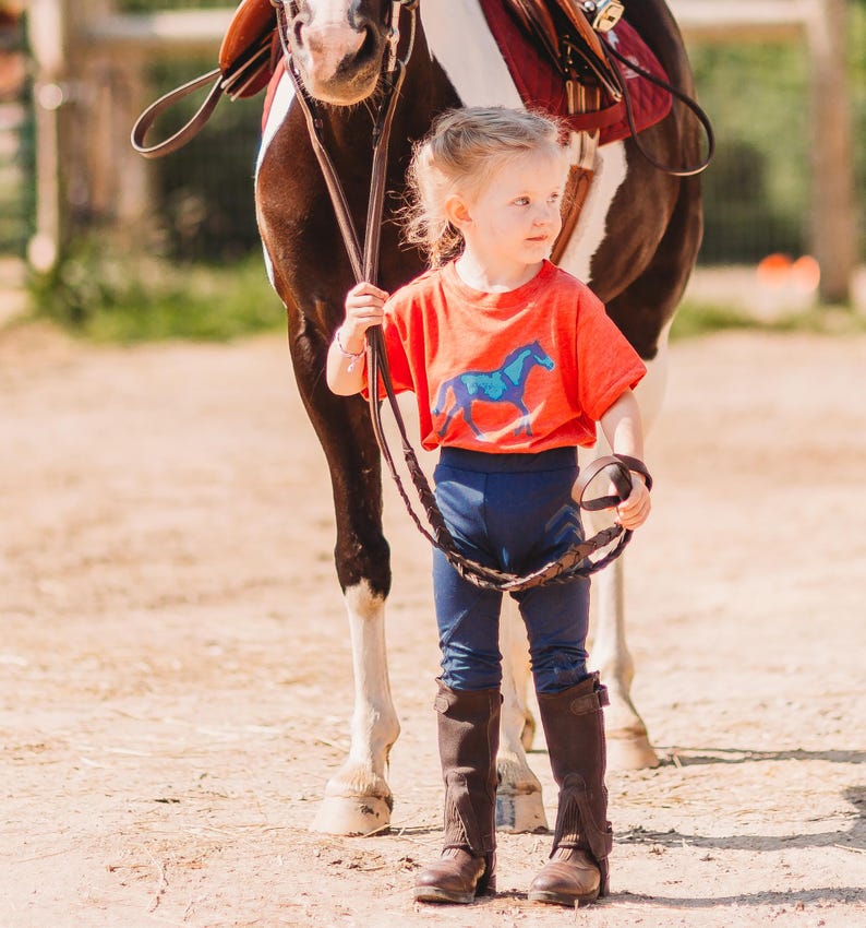 May include: A young child wearing an orange t-shirt with a blue horse graphic, navy blue leggings, and brown riding boots stands next to a horse. The horse has a brown and white coat and is wearing a saddle.