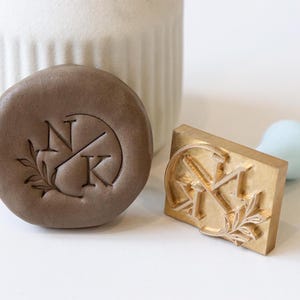 May include: A close-up of two brass stamps with a leaf design and the initials "NK". One stamp is brown and the other is gold.