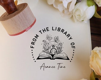 May include: A wooden stamp with a red ink pad. The stamp features a black and white illustration of a book with a house inside, surrounded by flowers and butterflies. The text "From the Library of..." and "Aimee Time" is printed around the illustration.