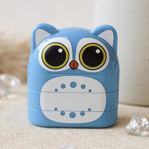 May include: A blue plastic owl-shaped pencil sharpener with large black eyes and a yellow iris. The sharpener has a white section with a row of small circles.