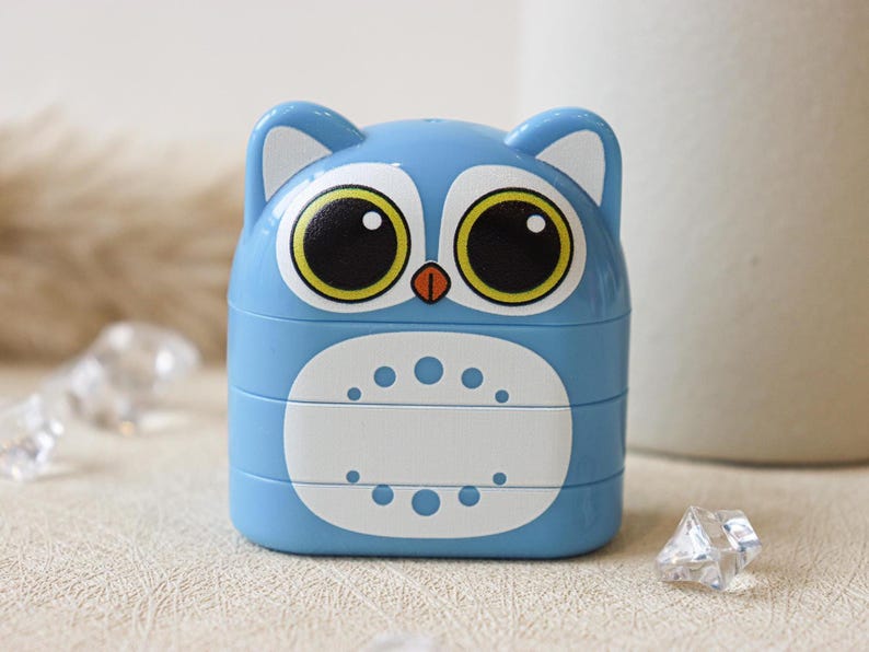 May include: A blue plastic owl-shaped pencil sharpener with large black eyes and a yellow iris. The sharpener has a white section with a row of small circles.