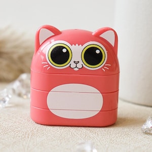 May include: A pink plastic cat-shaped container with three compartments. The cat has large yellow eyes and a white face.