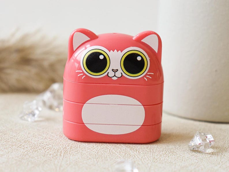 May include: A pink plastic cat-shaped container with three compartments. The cat has large yellow eyes and a white face.