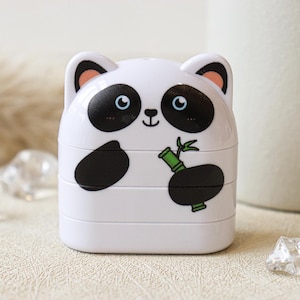 May include: A white plastic container shaped like a panda with black ears, eyes, and paws. The panda is holding a green bamboo shoot.