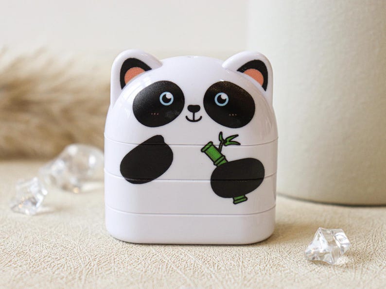 May include: A white plastic container shaped like a panda with black ears, eyes, and paws. The panda is holding a green bamboo shoot.