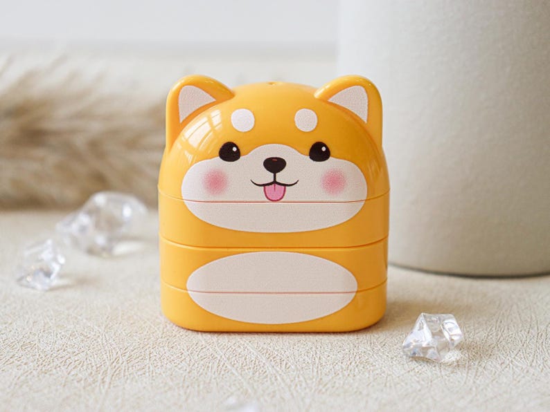 May include: A yellow plastic container shaped like a dog with a white face and pink cheeks. The container has a white oval on the bottom and is stacked on top of another container.