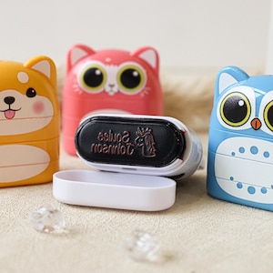 May include: Three colorful, stackable, self-inking stamps with animal designs. The stamps are yellow with a dog face, pink with a cat face, and blue with an owl face. The stamps are open, revealing the black ink pad with the text "Johnson Souvenirs" in red.