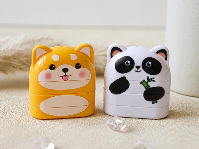 May include: Two small, stackable pencil sharpeners. One is yellow with a cartoon Shiba Inu face and the other is white with a cartoon panda face holding a bamboo shoot.
