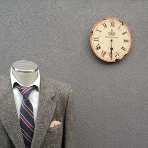 Puede incluir: Una chaqueta de tweed gris con una corbata a rayas azul oscuro y roja. La chaqueta está en un maniquí con una cabeza plateada. Un reloj vintage con números romanos está en la pared detrás del maniquí.