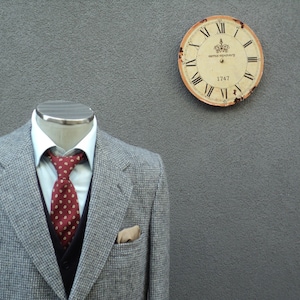 May include: A grey tweed blazer with a white shirt and a red patterned tie. The blazer has a pocket on the left side. A vintage clock with Roman numerals is on the wall behind the blazer.