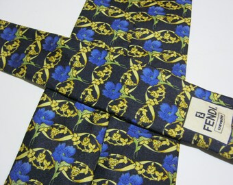 Puede incluir: Una corbata de seda negra con un patrón floral con flores azules y hojas amarillas. La corbata tiene una etiqueta FENDI con el texto "cravatte" impreso en ella.