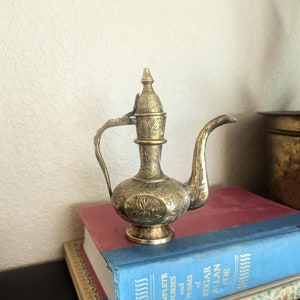 May include: A small, ornate brass teapot with a floral design. The teapot has a spout and a handle, and it sits on top of two books.