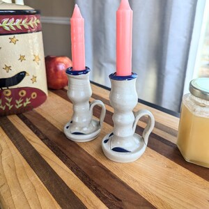May include: Two ceramic candle holders with blue and white glaze, each holding a pink candle. The candle holders are on a wooden surface.