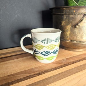 May include: White ceramic mug with a green and blue leaf pattern. The mug has a curved handle and a wide rim. The design features rows of stylized leaves in shades of green and blue. The mug is on a wooden surface.