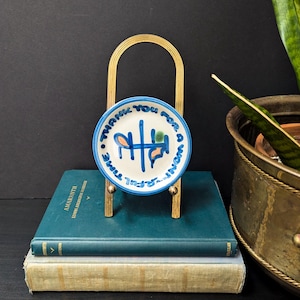 May include: A small, round ceramic plate with a blue rim and the words "THANK YOU FOR A WONDERFUL TIME" in blue lettering. The plate features abstract blue, orange, and green designs. It is displayed on a gold-colored metal stand, resting on a stack of books.