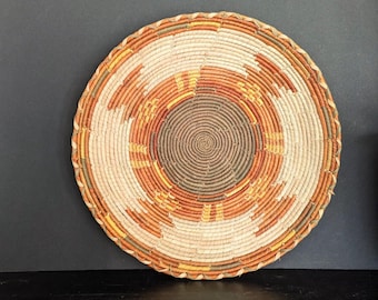 May include: A woven basket with a circular shape and a brown, white, and orange pattern. The basket is made of natural materials and has a traditional design.