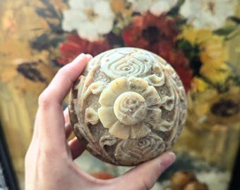 May include: A hand holds a round, carved, beige and brown stone box with a floral design. The box is intricately carved with a raised flower in the center and smaller flowers around the edges.