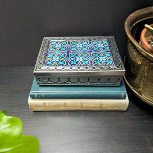 May include: A square metal box with a decorative tile lid featuring blue and yellow floral patterns. The box sits atop two stacked books. The box has a decorative border and is silver in color.