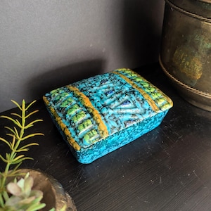 May include: A blue ceramic trinket box with a geometric pattern in green, yellow, and blue. The box has a speckled glaze and a rectangular shape.