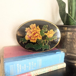 May include: An oval-shaped black lacquered trinket box with a floral design. The box is decorated with yellow and red flowers and green leaves. The box is sitting on top of two books.
