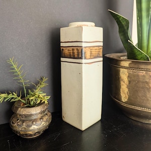 May include: A white ceramic vase with brown stripes. The vase has a square shape and a textured surface.