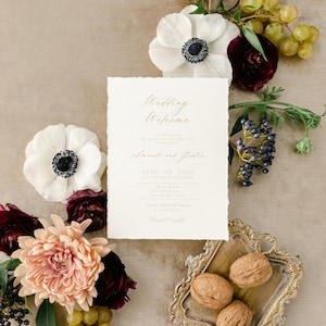 Op de afbeelding: Een welkomstsbord voor een bruiloft met witte bloemen, druiven en walnoten op een beige achtergrond. Het bord vermeldt "Wedding Welcome" en bevat de namen "Amanda en Justin", de datum "April 23, 2022", de locatie "Billy Bob's" en de dresscode "Casual Western Wear".