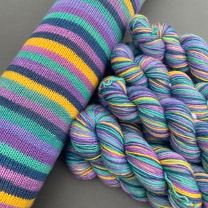 May include: A close-up of colorful yarn and a sock. The yarn is in skeins and the sock is rolled up. The colors include teal, purple, yellow, and navy blue. The yarn and sock are on a dark gray surface.