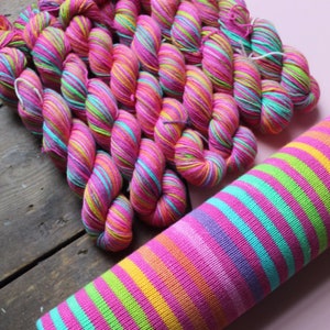 May include: A collection of vibrant, hand-dyed yarn skeins in shades of pink, turquoise, yellow, and green. The yarn is arranged on a wooden surface, with a knitted tube of the same color scheme in the foreground. The yarn is suitable for knitting and crochet projects.