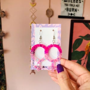 Puede incluir: Pendientes de brillo rosa y blanco con forma de símbolo femenino. Los pendientes están en una tarjeta con las palabras "back" e "itback" impresas en ella.