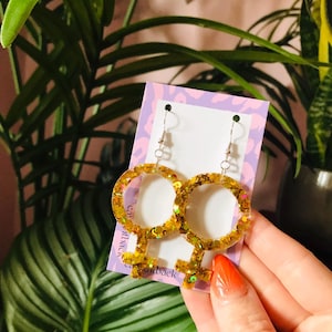 May include: A pair of gold glitter resin earrings in the shape of the female symbol. The earrings are suspended from silver-colored hooks. The background includes green plants and a purple card with text.