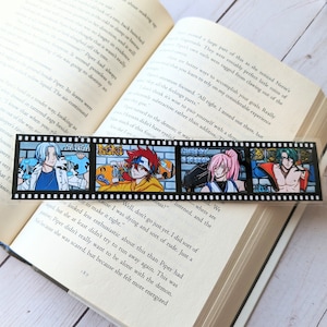 May include: A black and white film strip bookmark featuring four anime-style character illustrations. The bookmark is placed on an open book with printed text. The illustrations depict characters in various poses and outfits, with vibrant colors.