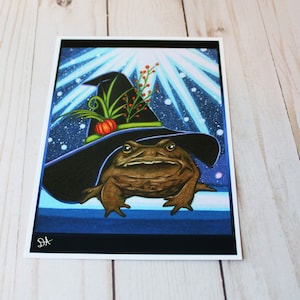 May include: A brown toad wearing a black witch's hat with a green brim and a small orange pumpkin on top. The hat is decorated with green leaves and red berries. The toad is sitting on a blue and white background with a starry sky and white snowflakes.