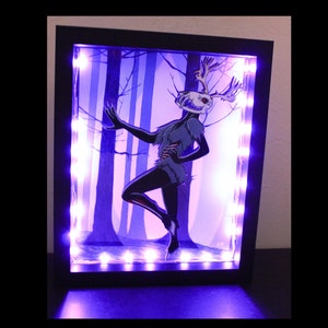 Puede incluir: Una ilustración enmarcada de una criatura de ciervo esquelético con cuernos, de pie en un entorno de bosque. El marco está iluminado con luces LED moradas.