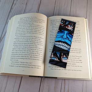 Pode incluir: Um marcador de livros preto com uma ilustra&ccedil;&atilde;o em estilo mang&aacute; de um rosto com olhos azuis e vermelhos e um rosto azul com dentes brancos. O marcador de livros est&aacute; em um livro aberto.
