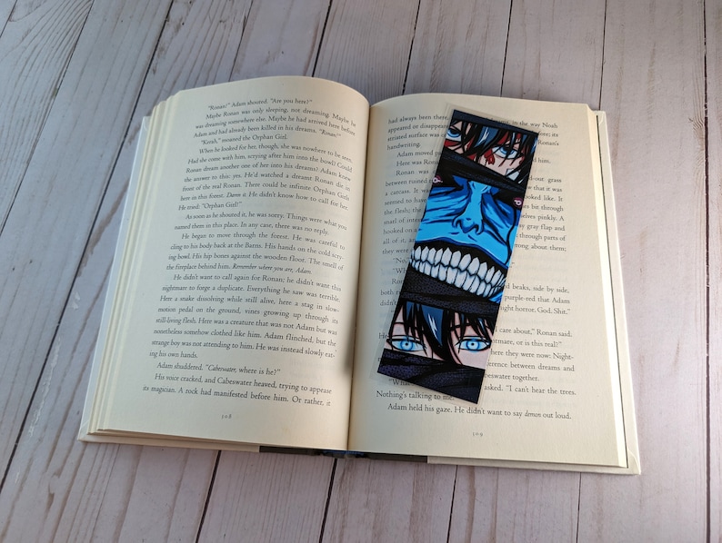 Pode incluir: Um marcador de livros preto com uma ilustra&ccedil;&atilde;o em estilo mang&aacute; de um rosto com olhos azuis e vermelhos e um rosto azul com dentes brancos. O marcador de livros est&aacute; em um livro aberto.
