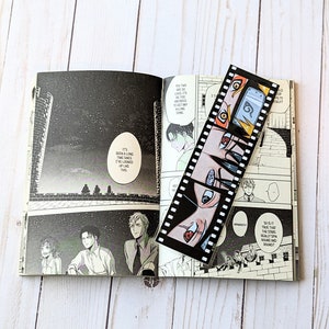 May include: A black and white manga bookmark with a film strip design featuring characters with red, blue, and orange eyes. The bookmark is laying open on a page of a manga book with a night sky background.