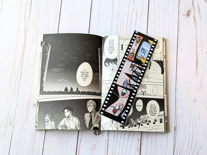 May include: A black and white manga bookmark with a film strip design featuring characters with red, blue, and orange eyes. The bookmark is laying open on a page of a manga book with a night sky background.