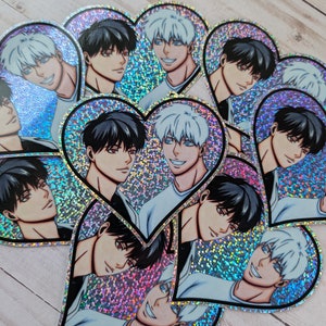 May include: A set of heart-shaped stickers featuring two anime characters. The characters have black and white hair and are depicted in a romantic pose. The stickers are holographic and have a glittery finish.