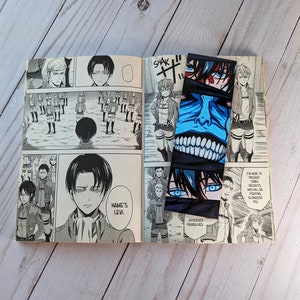 Pode incluir: Um livro aberto com ilustra&ccedil;&otilde;es de mang&aacute; e dois marcadores de Attack on Titan. Os marcadores apresentam arte de personagens com detalhes em azul e preto. As p&aacute;ginas do livro exibem pain&eacute;is de mang&aacute; em preto e branco.