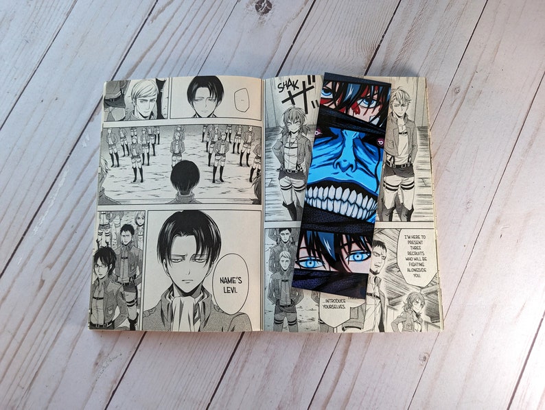 Pode incluir: Um livro aberto com ilustra&ccedil;&otilde;es de mang&aacute; e dois marcadores de Attack on Titan. Os marcadores apresentam arte de personagens com detalhes em azul e preto. As p&aacute;ginas do livro exibem pain&eacute;is de mang&aacute; em preto e branco.
