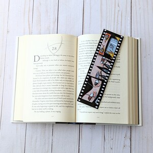 May include: A black and white bookmark with a film strip design featuring a group of anime characters with black hair and colorful eyes. The bookmark is placed in an open book with white pages.
