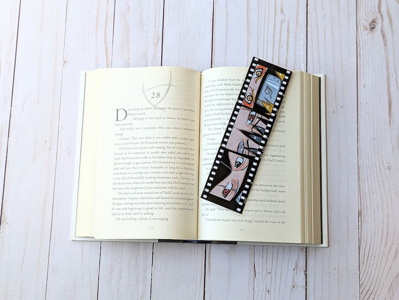 May include: A black and white bookmark with a film strip design featuring a group of anime characters with black hair and colorful eyes. The bookmark is placed in an open book with white pages.