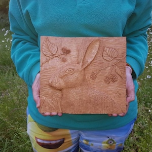 May include: A wood carving of a rabbit's head with a branch of leaves and berries. The carving is on a square piece of wood and is signed "J.L. 2017".