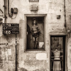 May include: A sepia photograph of a building facade featuring a statue in a recessed window. The sign above reads "VIA DEGLI ALFANI." A shop doorway reflects a dress form. Directional signs point to local landmarks.