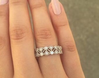 May include: A silver ring with a repeating pattern of diamonds and small round gemstones. The ring is on a person's finger, with a light pink nail polish. The ring's design features a series of diamond-shaped cutouts and small, round gemstones.