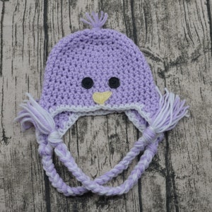 May include: Purple crocheted hat with white and purple braided ties. The hat has a bird design with black button eyes and a yellow beak.