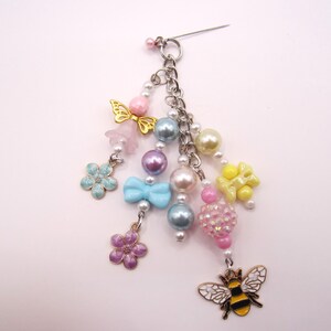 May include: A decorative brooch featuring a silver-tone pin base with multiple dangling charms including a bee, flowers, a bow, and various colorful beads in shades of pink, blue, yellow, and purple, arranged on silver-tone metal chains.