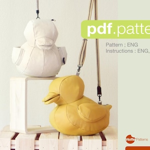 Op de afbeelding: Twee eendvormige tassen, één crèmekleurige en één gele, worden getoond. De gele tas heeft een riem. De tassen staan op een houten oppervlak. De afbeelding bevat de tekst "pdf.pattern" en "Pattern : ENG Instructions : ENG, JP".