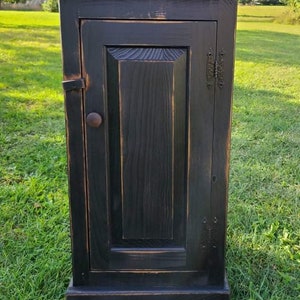 May include: A small, black wooden cabinet with a single door. The cabinet has a distressed finish, revealing hints of the wood grain. It features a knob and hinges, and sits on a base. The cabinet is outdoors on a grassy area.